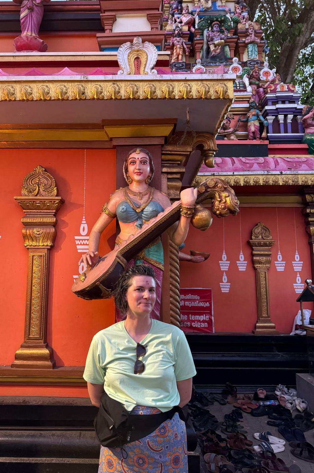 A person is standing in front of a colorful temple with intricate carvings, including a prominent statue of a figure holding a musical instrument.